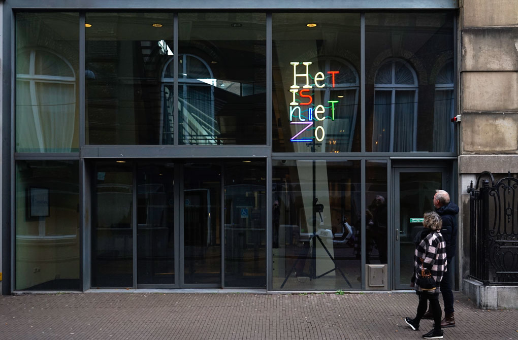 In gekleurde neonletters op een vier meter hoog statief staat Het is niet zo. Het kunstwerk staat bij ingang van het Logement van de Heeren van Amsterdam. Het is het gebouw waar de Parlementaire Enquête Commissie van de Tweede Kamer zitting heeft. Het kunstwerk is gefotografeerd na de restauratie op 13 december 2022