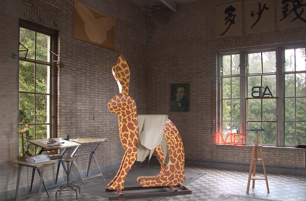 interieur van het atelier met o.a. de Giraf, het beeld is in cortenstaal uitgevoerd en staat in Belgie, Beerse. Links boven aan de muur een papieren vlindervorm van twee meter lang en een meter breed. 
        Deze vorm is in 120 mm dik zwart graniet uitgehakt en gepolijst, © André Boone