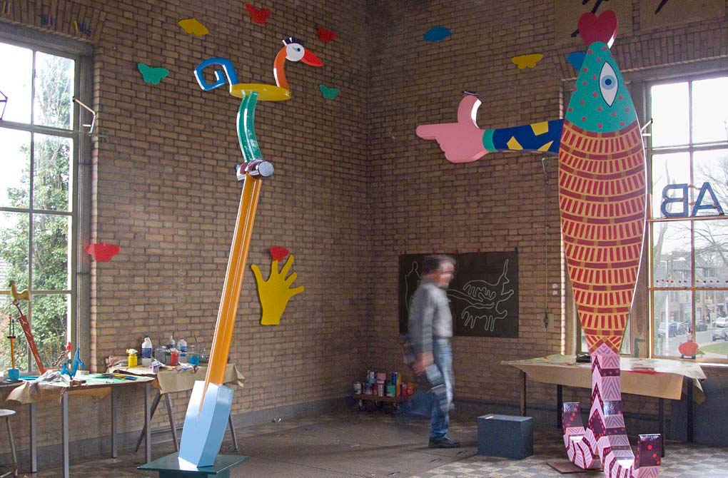 atelier met op de voorgrond een gekleurde cortenstalen vogel op een schep, aan de muur een gele hand met een geamputeerde vinger. Op een wijsvinger zit een rode vlinder. Rechts een verticale vis met een arm waarvan de duim omhoog steekt, thumbs up. Rechts op de muur een ontwerp, een boer achter een eergetouw voortgetrokken door twee runderen. In de hoek potten met verf, 20 kleuren, werden gebruikt om het beeld Daar was laatst een meisje loos te schilderen.  Op de rechter vensterbank staat een egel met een ster op zijn rug.op de linkervensterbank staan een aantal schaalmodellen