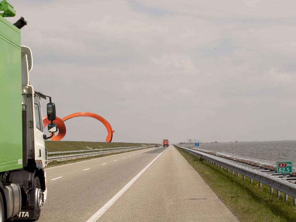 een montage, het monumentale ontwerp op de afsluitdijk verdwijnt gedeelte  achter een rijdende vrachtauto, © André Boone