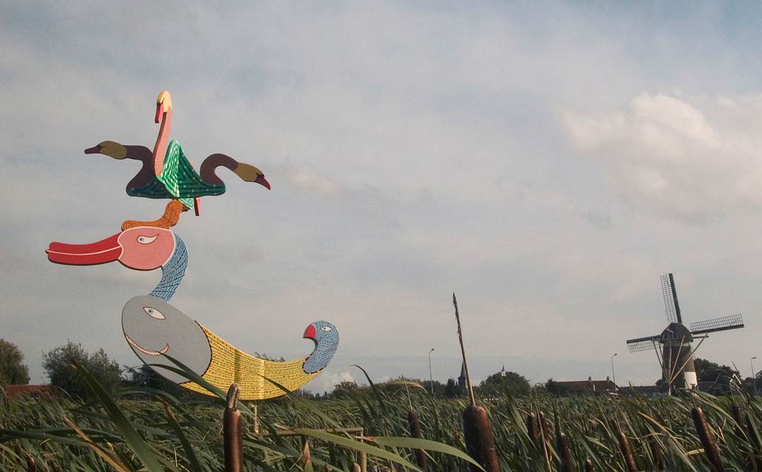 een windobject draaiend op drie punt. Het staat in het riet in de polder. het is een grote eend met op zijn kuif vier zwamen halsen met kop, © André Boone